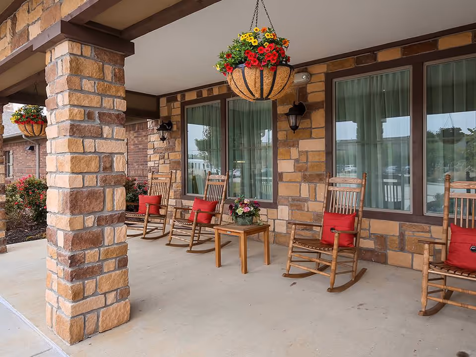 Covered outdoor patio area with stone pillars and a stone wall. There are four wooden rocking chairs with red cushions arranged around a small wooden table with a flower arrangement. Hanging flower baskets with red and yellow flowers are suspended from the ceiling.
