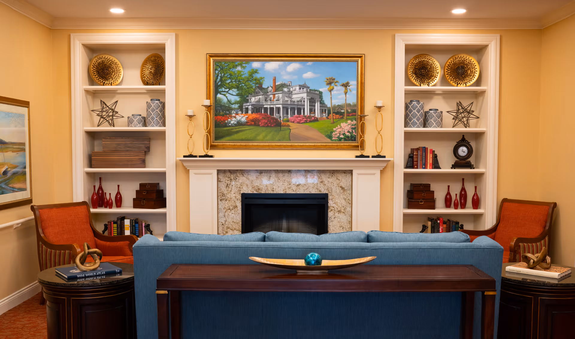 Cozy living room with a blue sofa facing a fireplace. On either side of the fireplace are white built-in shelves decorated with books, vases, and decorative objects. Two orange armchairs flank the sofa, and a framed painting of a large house with a garden hangs above the fireplace.