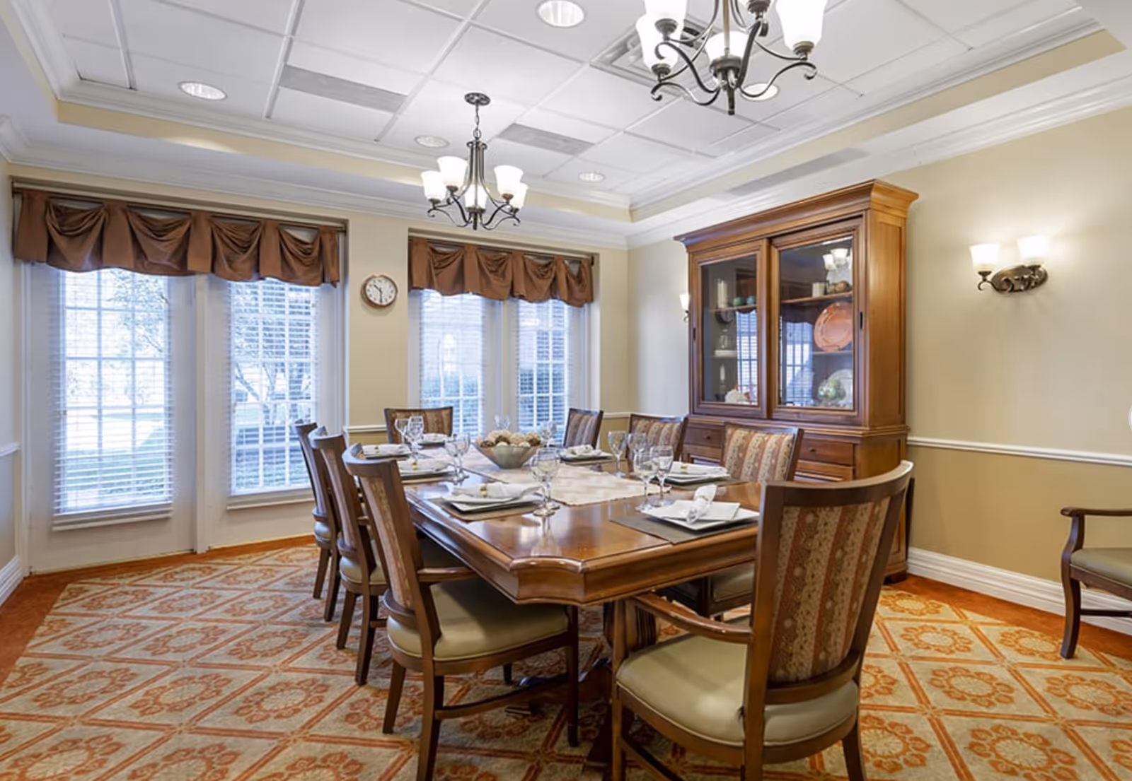 Formal dining room with a long wooden table set for a meal, upholstered chairs, a china cabinet, chandeliers and large windows with valances.