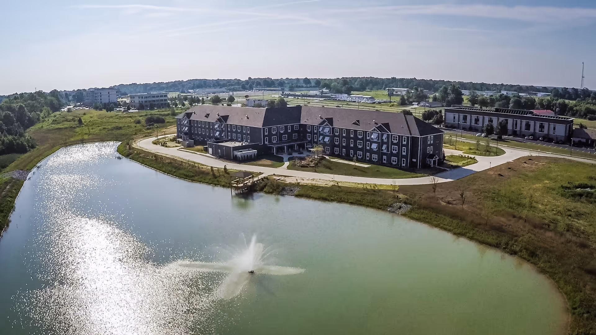 Aerial view of The Landings at Reed Station Crossing, showing a large building complex next to a pond with a water fountain. The surrounding area includes greenery, roads, and additional buildings in the distance under a clear sky.