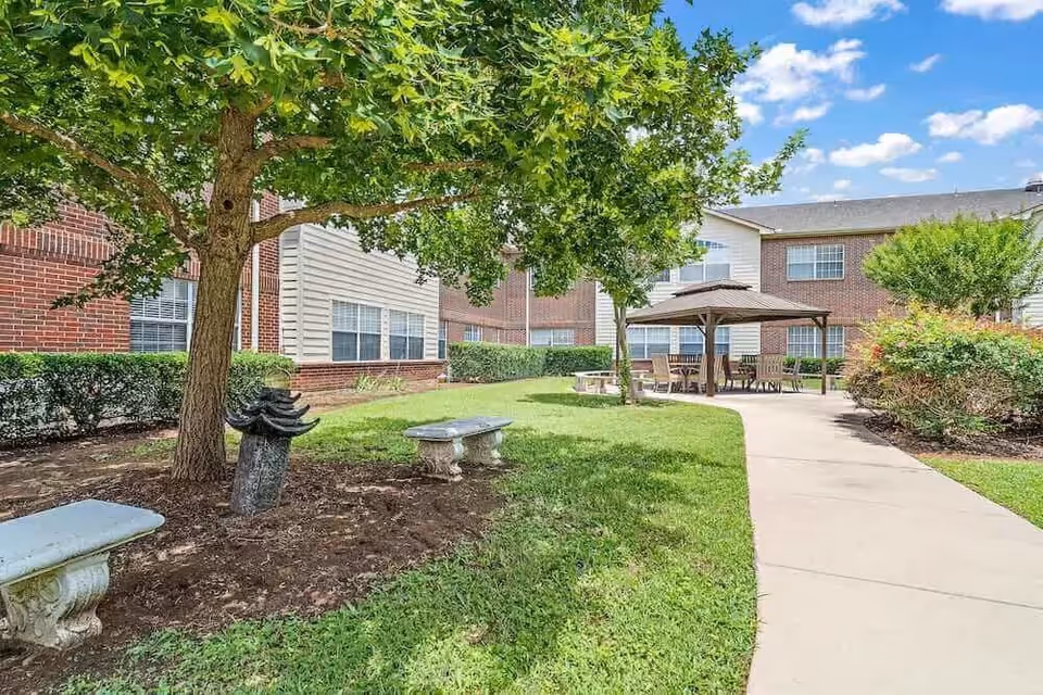 Outdoor courtyard area at Morada Lake Arlington featuring a paved walkway, green grass, trees, two stone benches, and a covered seating area with chairs. The background shows a two-story brick and siding building under a blue sky with some clouds.