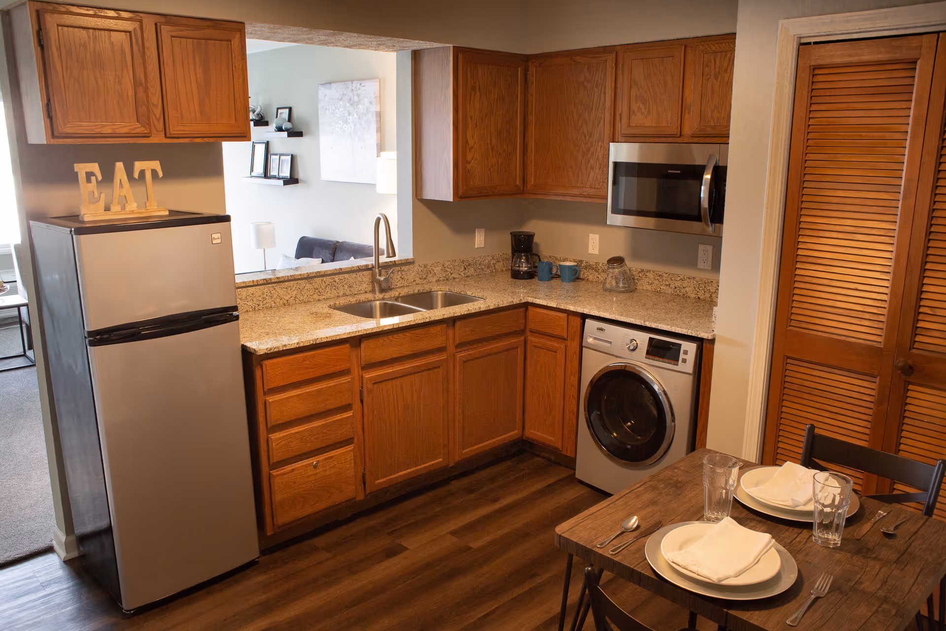 Compact kitchen with oak cabinets, granite countertops, stainless refrigerator and microwave, a washing machine under the counter, and a small dining table set for two.