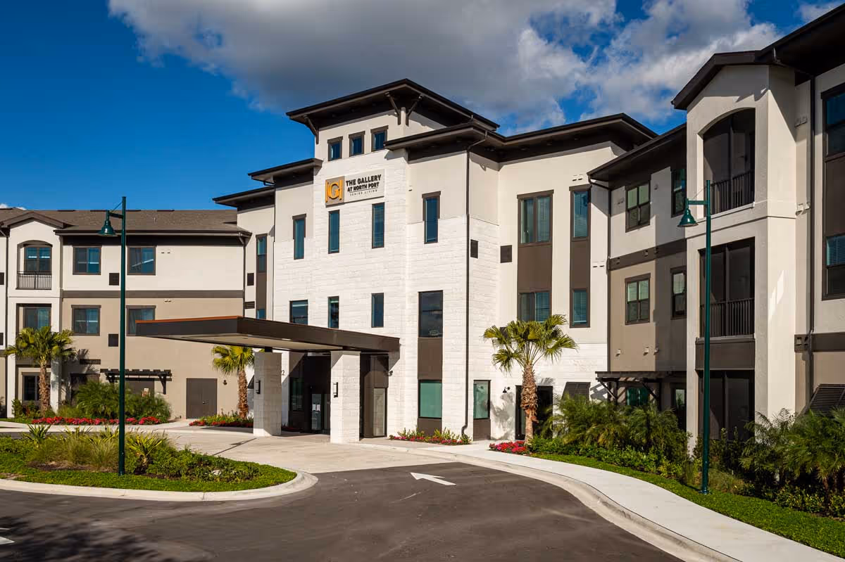 Exterior view of The Gallery at North Port, a modern multi-story senior living facility with beige and white walls, large windows, palm trees, and a covered entrance under a blue sky with some clouds.
