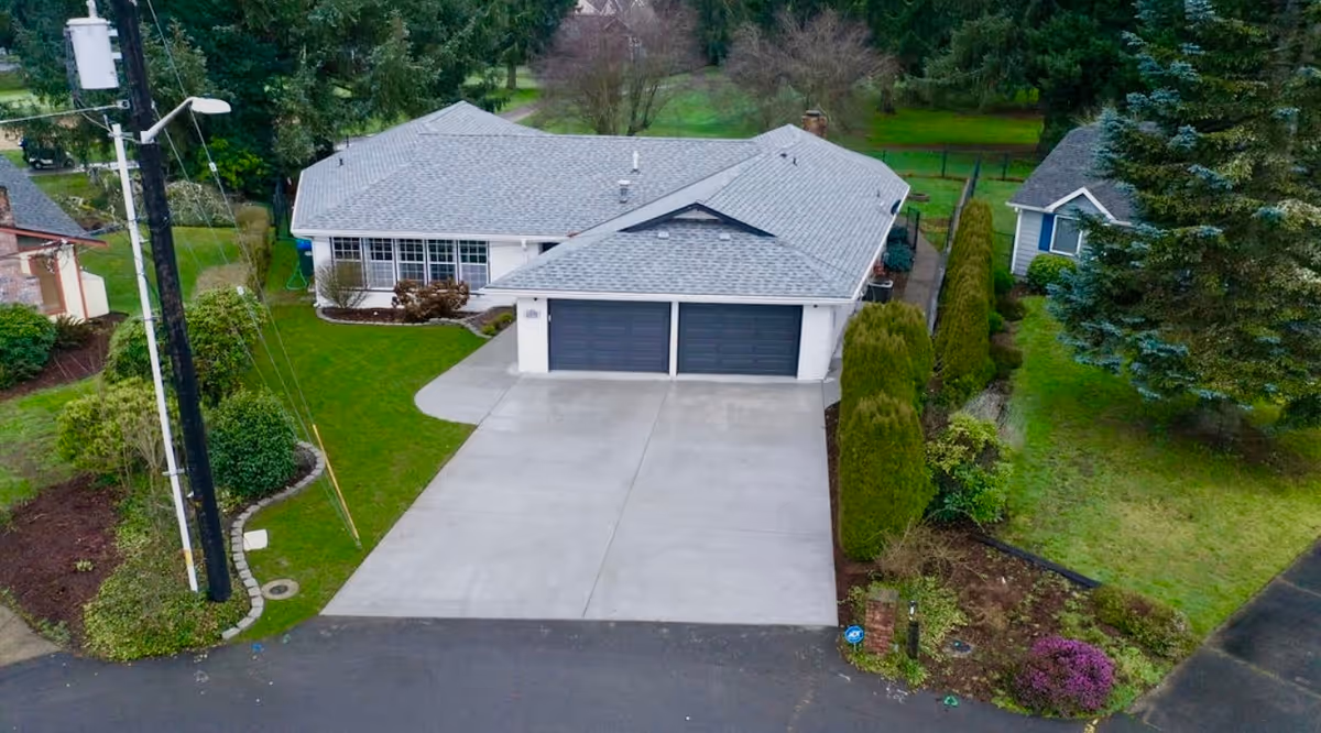 Aerial view of a single-story residential building with a gray roof and two-car garage. The house is surrounded by green lawns, shrubs, and trees. There is a paved driveway leading to the garage and neighboring houses visible on either side.