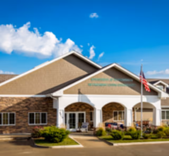 Front exterior of Edencrest at Riverwoods with arched entrance and American flag under blue sky.