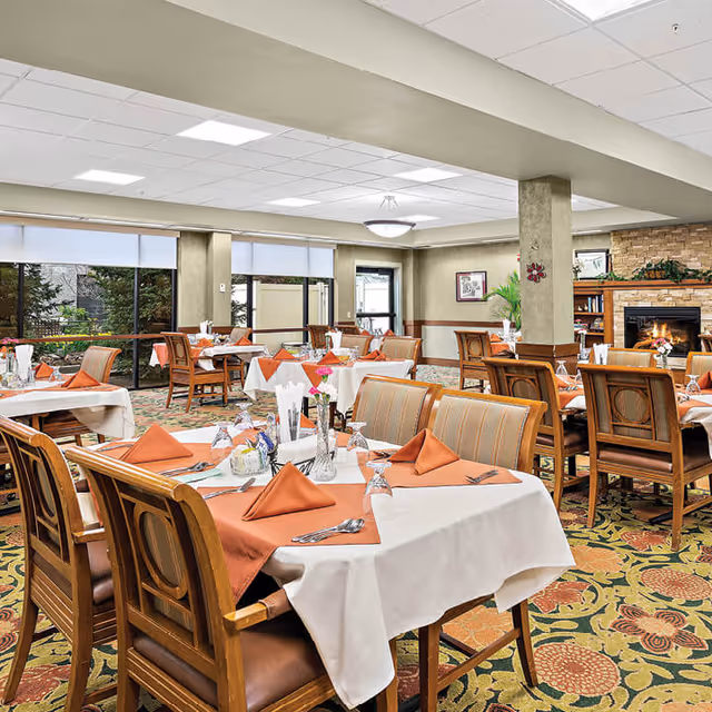 A dining room with multiple tables covered in white tablecloths and orange napkins, set with glasses, silverware, and floral centerpieces. The room has large windows letting in natural light, a patterned carpet, wooden chairs, and a stone fireplace in the background.