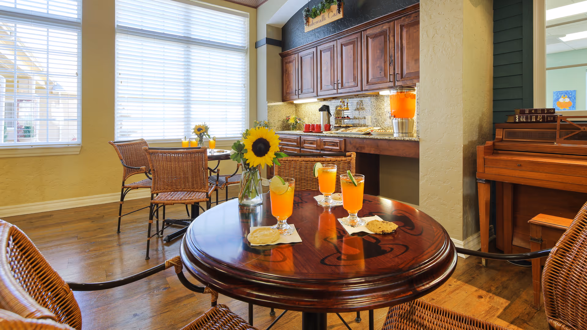 A cozy dining area with wooden floors and wicker chairs around round wooden tables. On the nearest table, there are three glasses of orange drink garnished with lime slices, a vase with a sunflower, and some cookies on napkins. In the background, there is a counter with wooden cabinets, a beverage dispenser with orange drink, and a piano with books on top. Large windows with white blinds let in natural light.