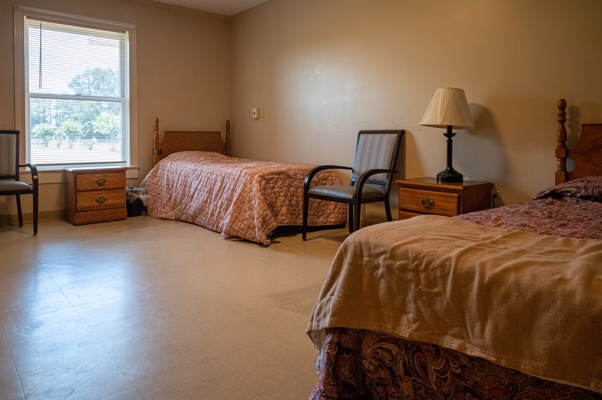 A simple senior living bedroom with two twin beds, wooden nightstands, chairs, a lamp, and a window.