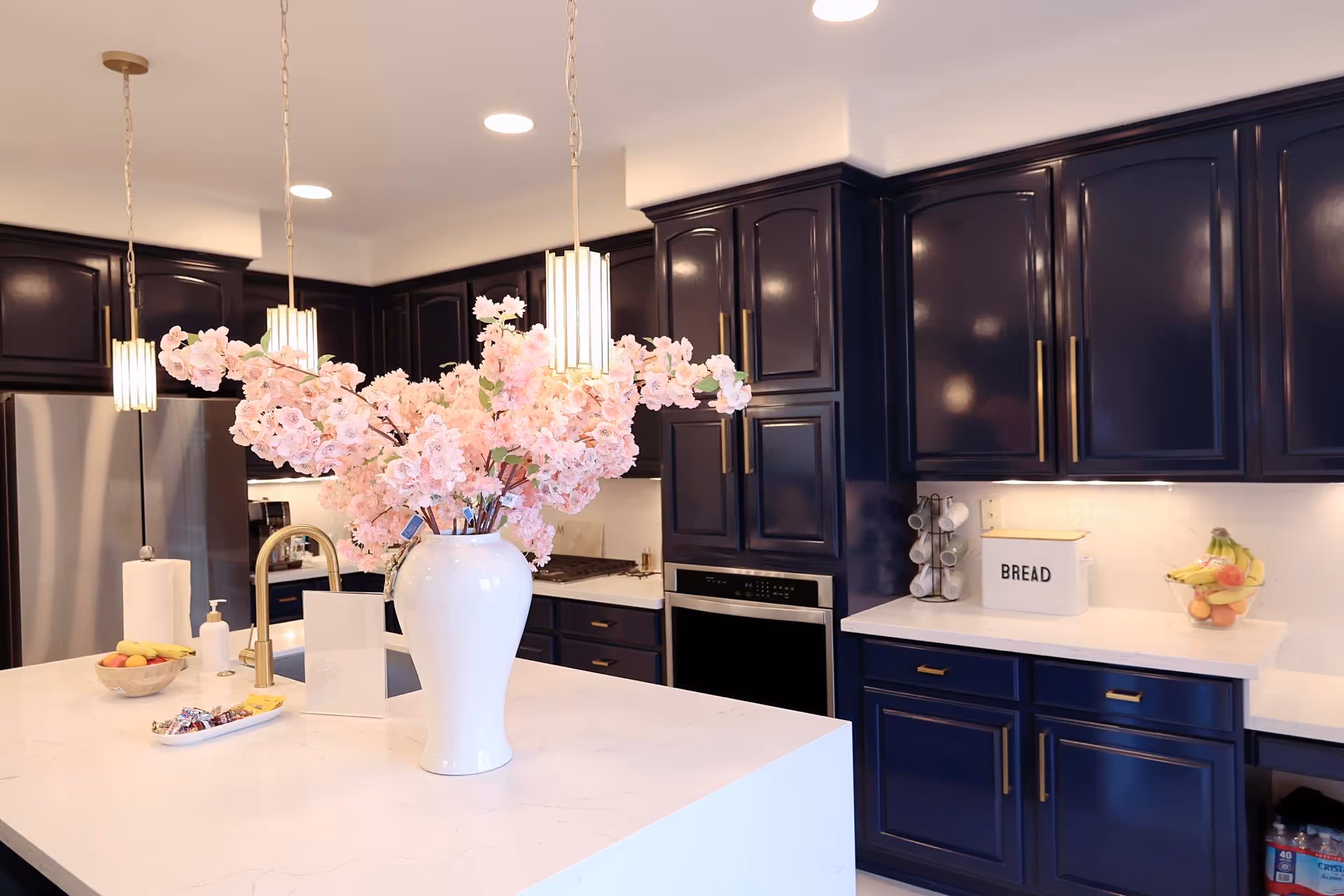 Modern kitchen with dark blue cabinets, white marble countertops, and a large white island in the center. The island features a white vase with pink flowers, a gold faucet, a bowl of fruit, and a tray with snacks. Pendant lights hang above the island, and stainless steel appliances are visible in the background.