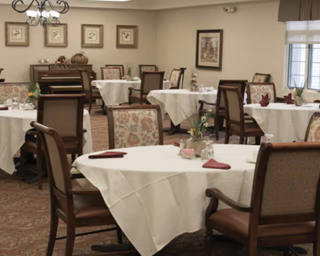 Dining room with round tables covered in white tablecloths, each set with folded burgundy napkins, glassware, and small floral centerpieces. The room has upholstered chairs with floral patterns and a piano against the far wall, decorated with small seasonal items. Framed artwork hangs on the walls, and a window with a valance allows natural light into the space.