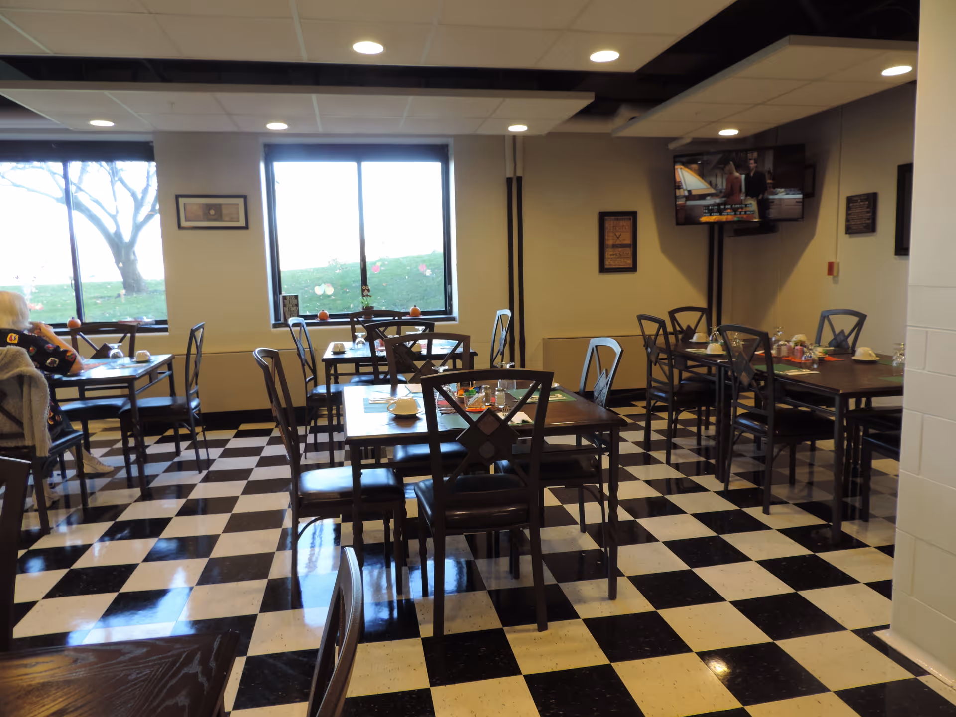 Dining room with black-and-white checkered floor, multiple tables and chairs set for meals and windows overlooking a lawn.