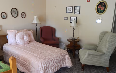 Cozy bedroom with a single bed dressed in pink bedding, two upholstered armchairs, side tables with lamps, and framed pictures on the wall.