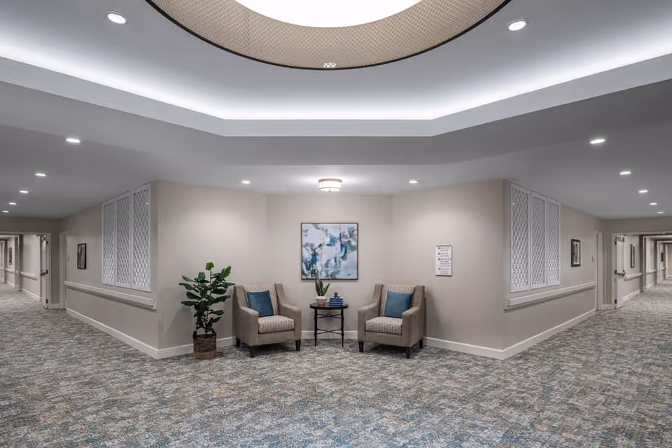 A well-lit hallway corner in a senior living facility with two beige armchairs featuring blue cushions, a small round table with a plant and books between them, a potted plant to the left, abstract artwork on the wall, and patterned carpet flooring extending down two corridors.