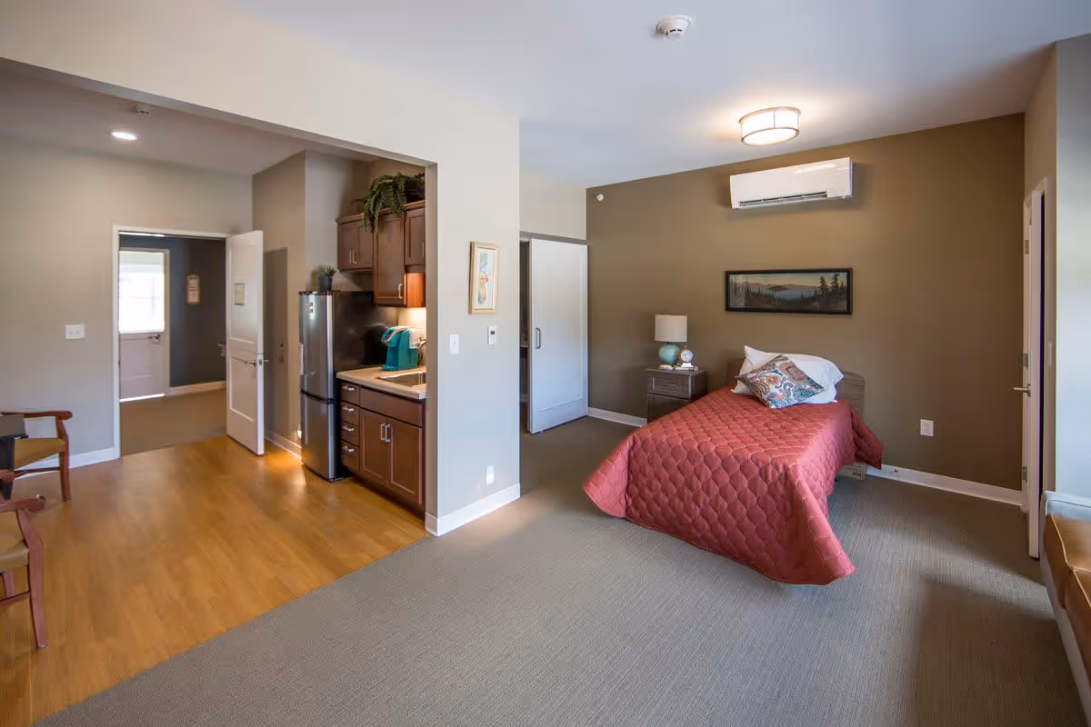 A cozy room in a senior living facility featuring a single bed with a red quilt and decorative pillows, a nightstand with a lamp, a small kitchenette with a refrigerator and coffee maker, and a seating area with chairs. The room has a warm color palette with beige and brown walls, wood flooring in the kitchenette area, and carpet in the sleeping area.