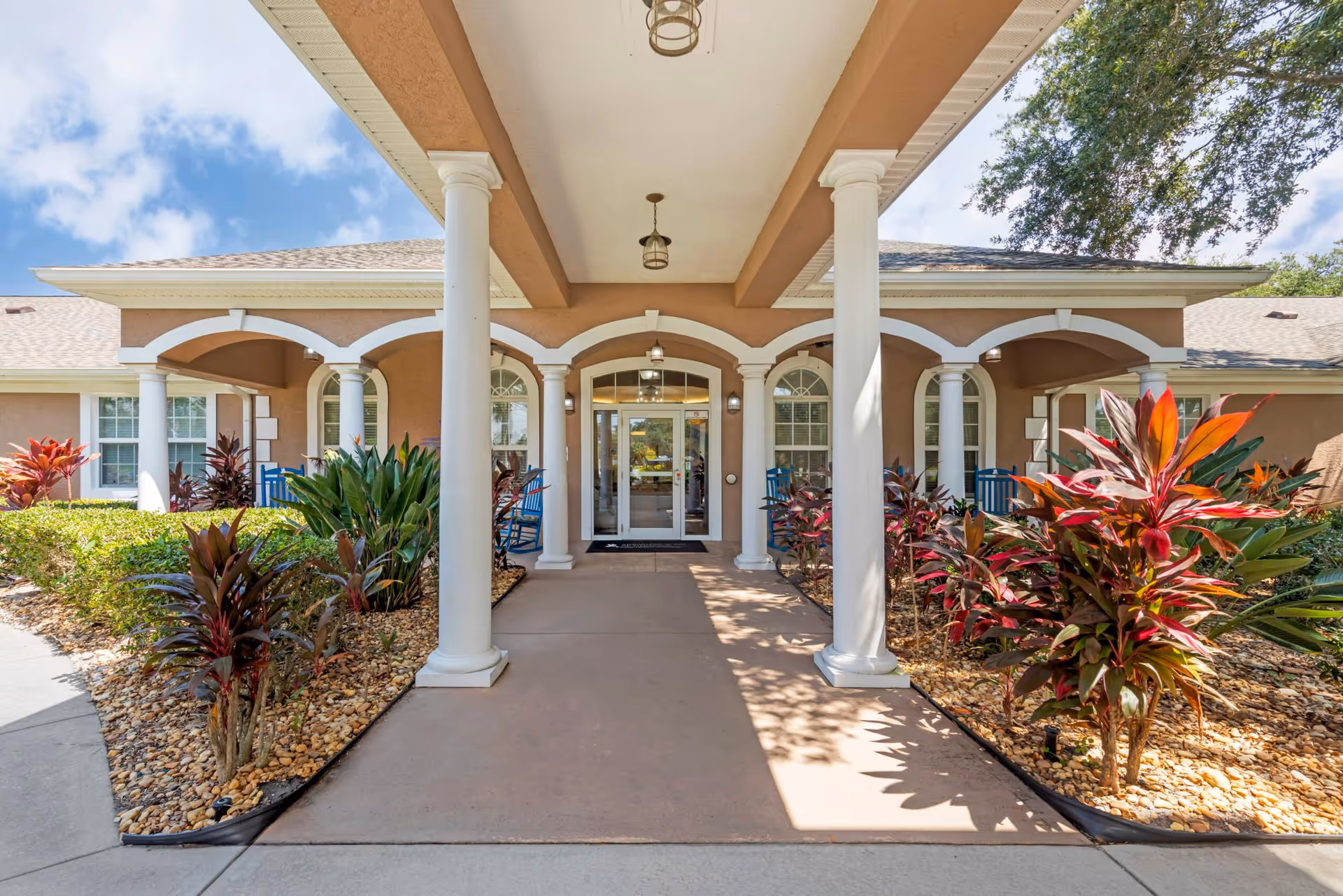Covered entrance of a senior living building with white columns, landscaped plantings, and glass double doors.