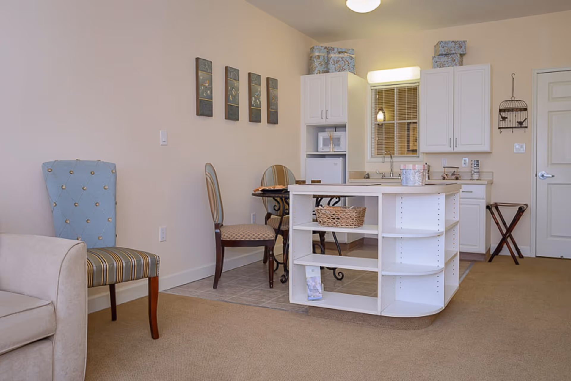 Interior view of a small living space featuring a beige armchair and a blue striped chair on the left, a small dining table with two chairs in the middle, and a compact kitchen area with white cabinets, a small refrigerator, microwave, and a white island with open shelves. The walls are light-colored with four decorative wall hangings and some decorative boxes on top of the cabinets.