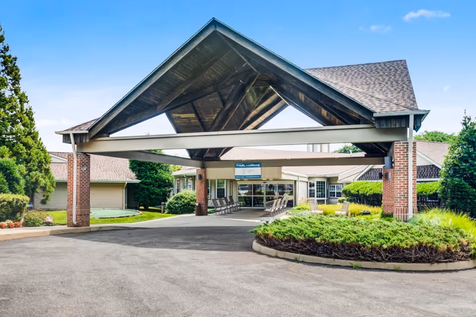 Entrance of Truewood by Merrill, Glen Riddle featuring a covered driveway with a peaked roof supported by brick pillars, surrounded by greenery and outdoor seating.