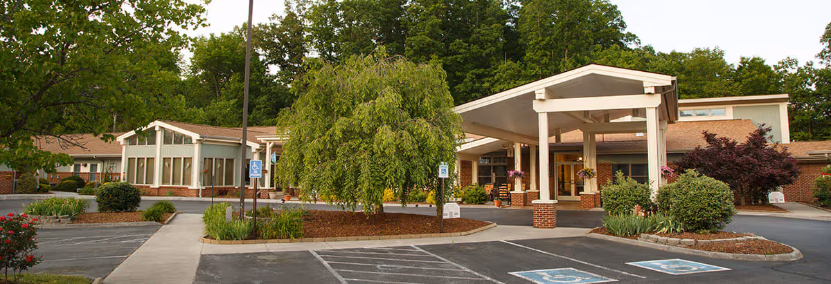 Exterior view of Preston Place Suites facility showing a single-story building with a covered entrance, surrounded by landscaped greenery and trees. There are parking spaces, including handicapped spots, in front of the building.