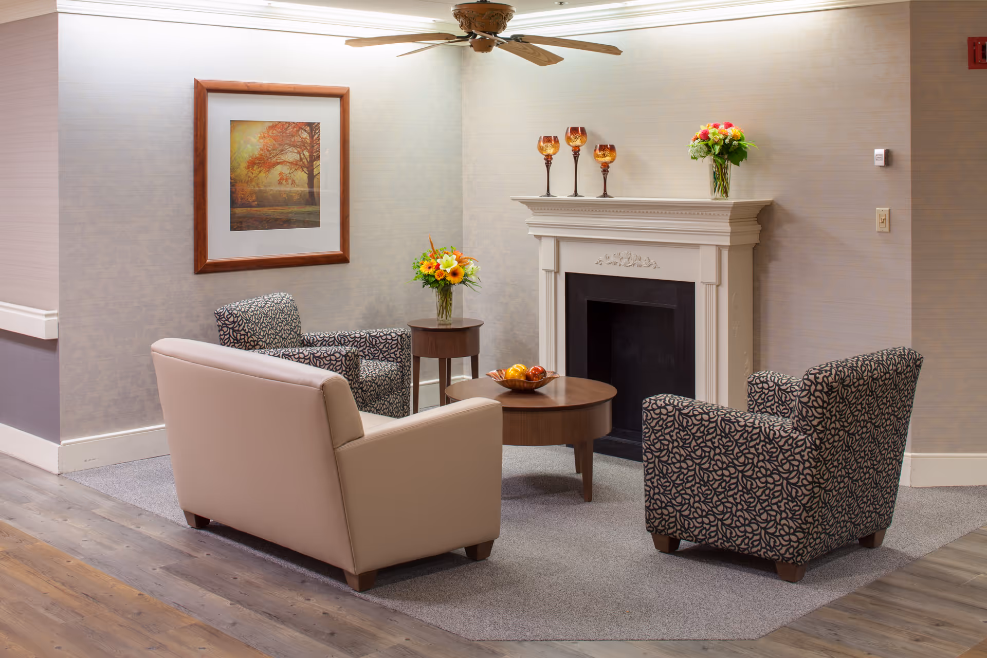 A cozy sitting area with a beige loveseat and two patterned armchairs arranged around a wooden coffee table with a bowl of fruit. Behind the seating is a white decorative fireplace mantel adorned with three amber-colored goblets and a vase of flowers. A framed picture of a tree with autumn leaves hangs on the wall, and a ceiling fan is visible above.