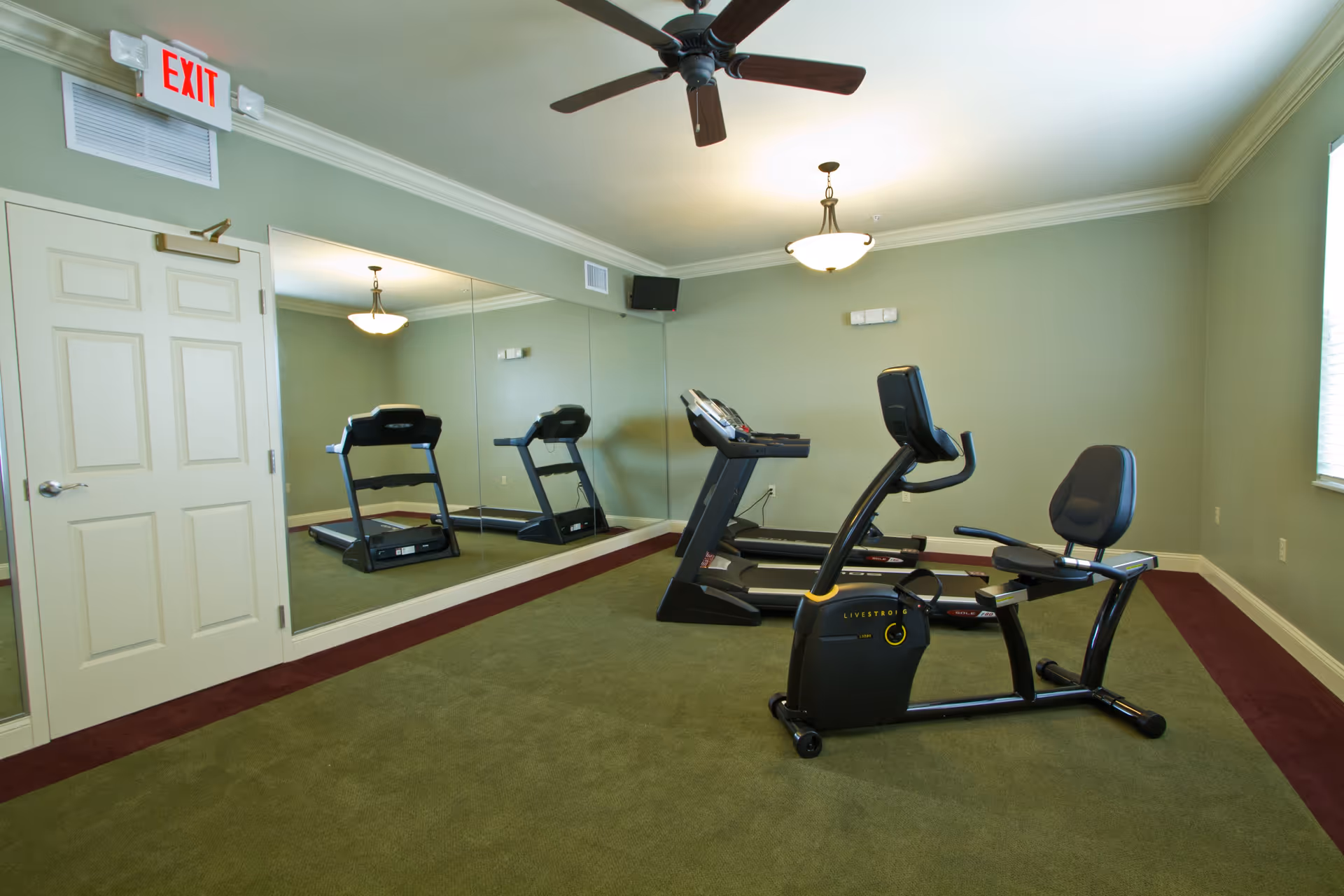 A small fitness room with green carpet and light green walls. The room contains two treadmills and a stationary recumbent bike. There is a large wall mirror reflecting the equipment, a ceiling fan, a hanging light fixture, and an exit door with an illuminated exit sign above it.