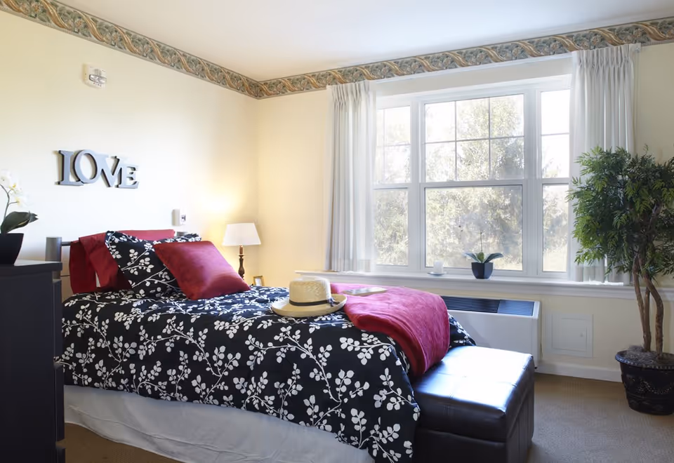 A bright bedroom with a large window letting in natural light. The bed has black and white floral bedding with red pillows and a red throw blanket. A straw hat rests on the bed. There is a white lamp on a nightstand beside the bed, a potted plant on the windowsill, and a larger potted plant in the corner. The wall has a decorative border near the ceiling and a wall decoration spelling 'LOVE'.