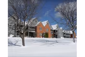 A row of multi-story residential buildings with brick and siding exteriors, surrounded by snow-covered ground and leafless trees under a clear blue sky.