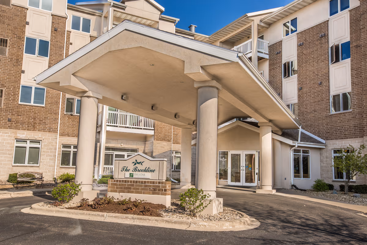 Entrance of a senior living facility named The Brookline with a covered driveway supported by large columns. The building is multi-story with brick and light-colored exterior walls, several windows, and a glass door entrance.