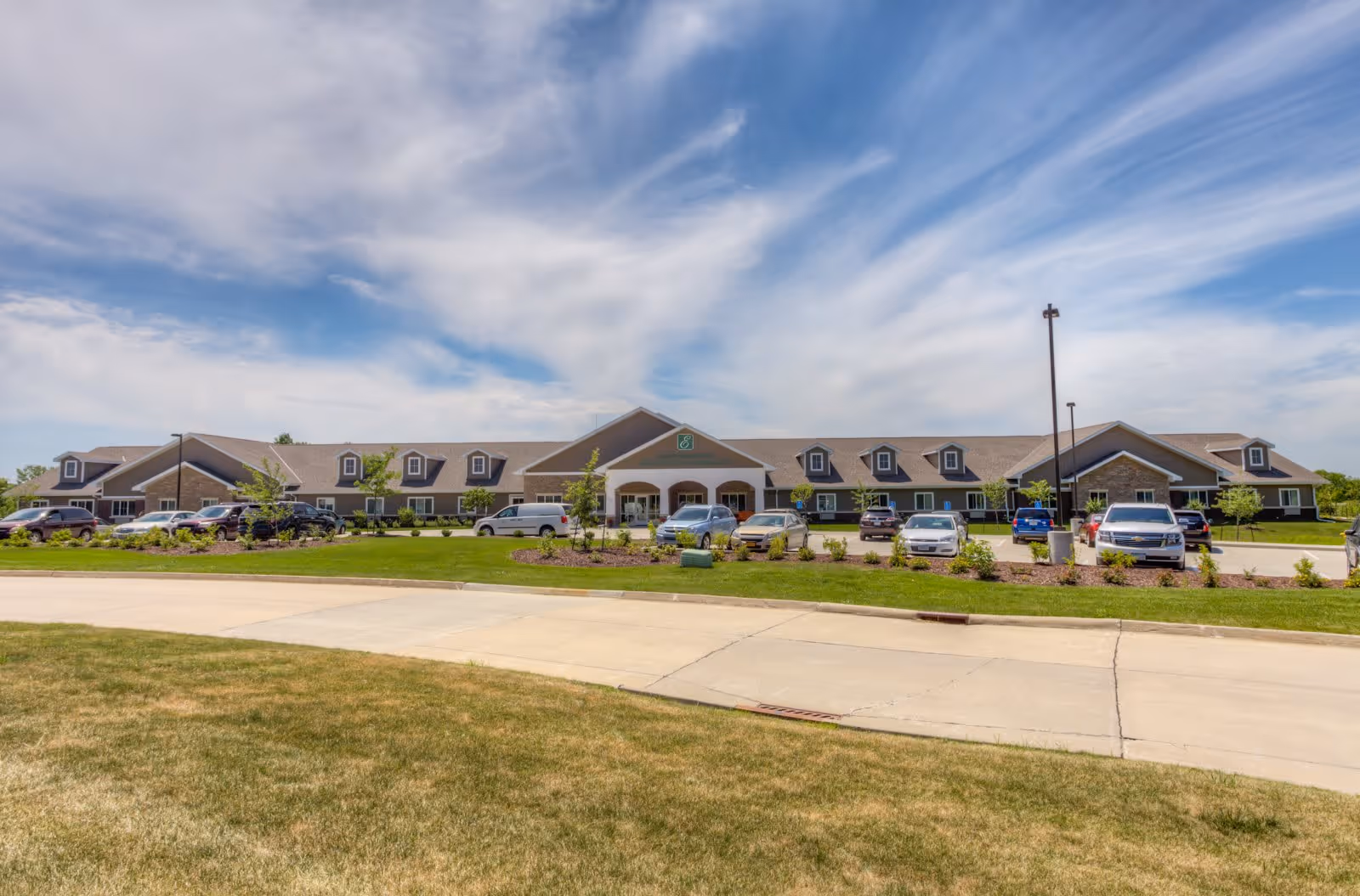 Wide exterior view of a single-story senior living facility building with multiple dormer windows and a central entrance. Several cars are parked in front of the building, and the area is landscaped with grass, small trees, and shrubs under a partly cloudy blue sky.