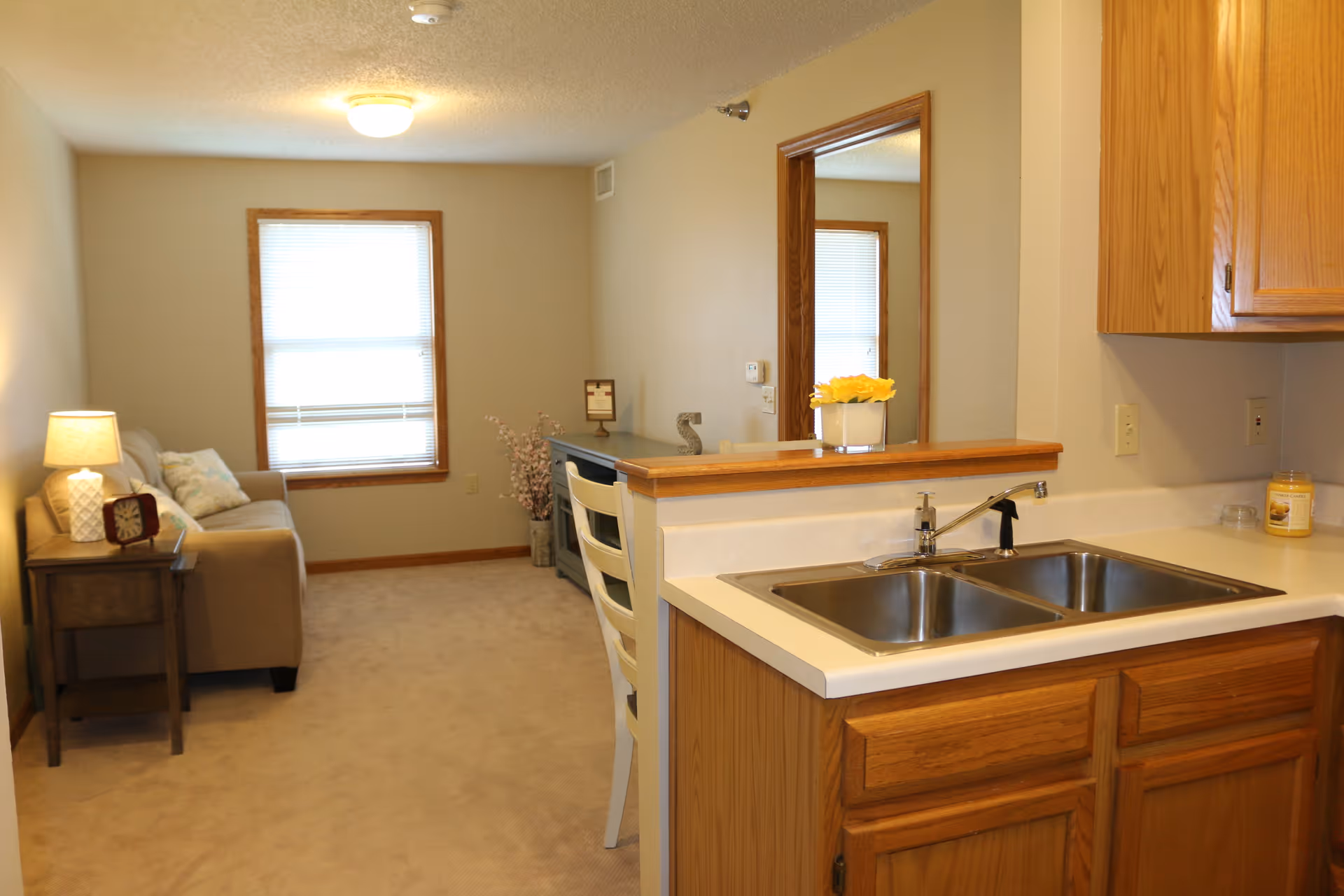 Interior view of a senior living facility showing a small living room area with a beige sofa, side table with a lamp and clock, a window with blinds, and a kitchen counter with a double sink and wooden cabinets. There is a mirror on the wall reflecting part of the room and a small vase with yellow flowers on the counter.