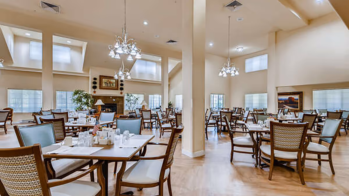 A spacious dining room with multiple wooden tables and chairs arranged neatly. The room has high ceilings with hanging chandeliers, large windows with white blinds, and a fireplace with framed artwork above it. The floor is light wood, and there are some potted plants near the fireplace.