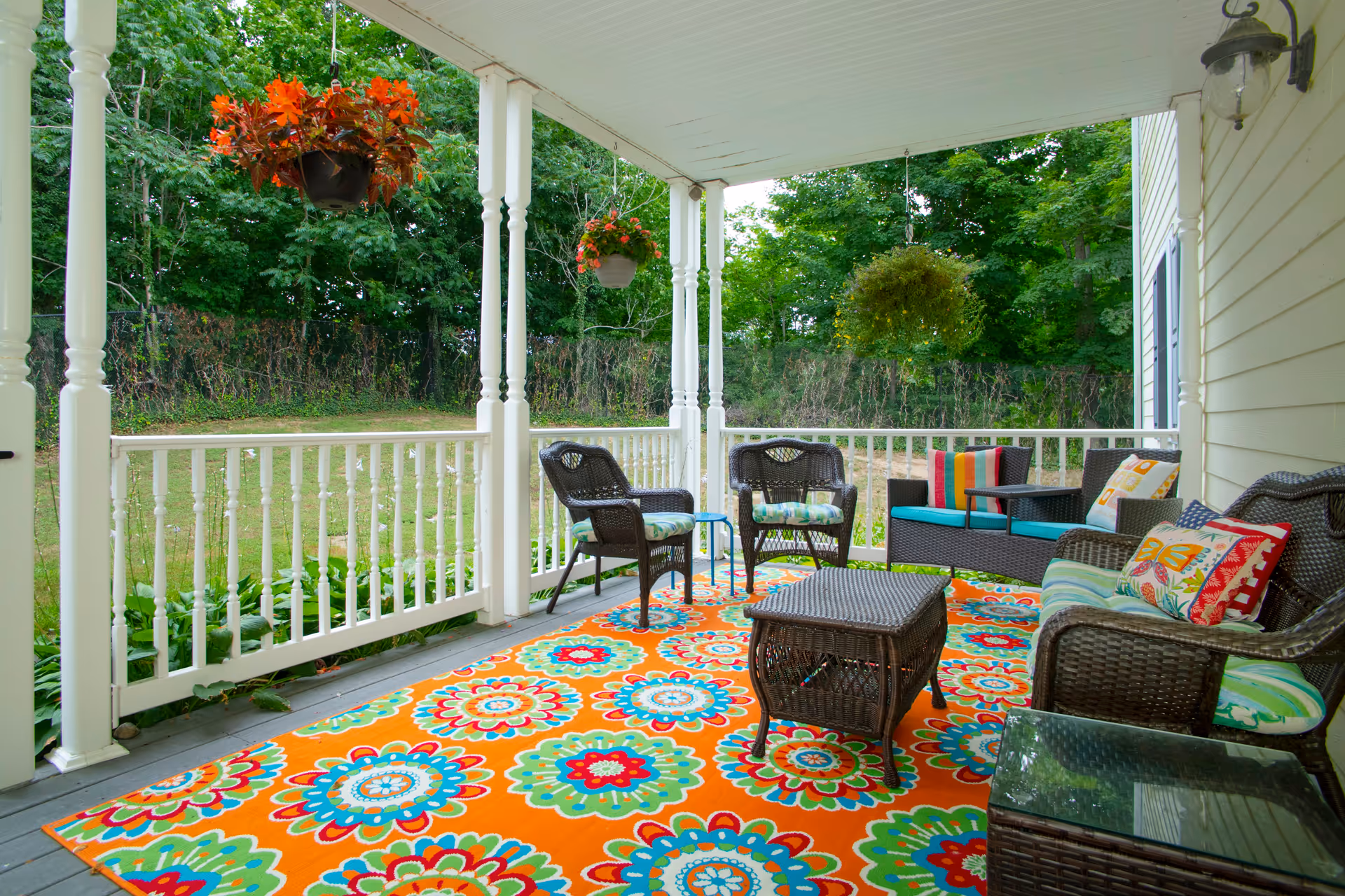 Covered porch with a bright floral rug, wicker seating, cushions, and hanging plants overlooking a grassy yard and trees.
