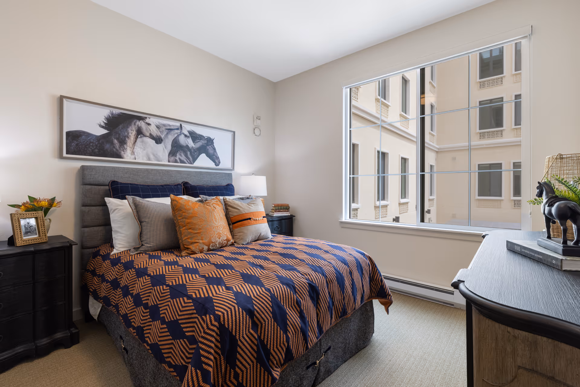 Furnished bedroom with a patterned bedspread, multiple pillows, bedside tables, horse artwork above the headboard, and a large window facing an interior courtyard.