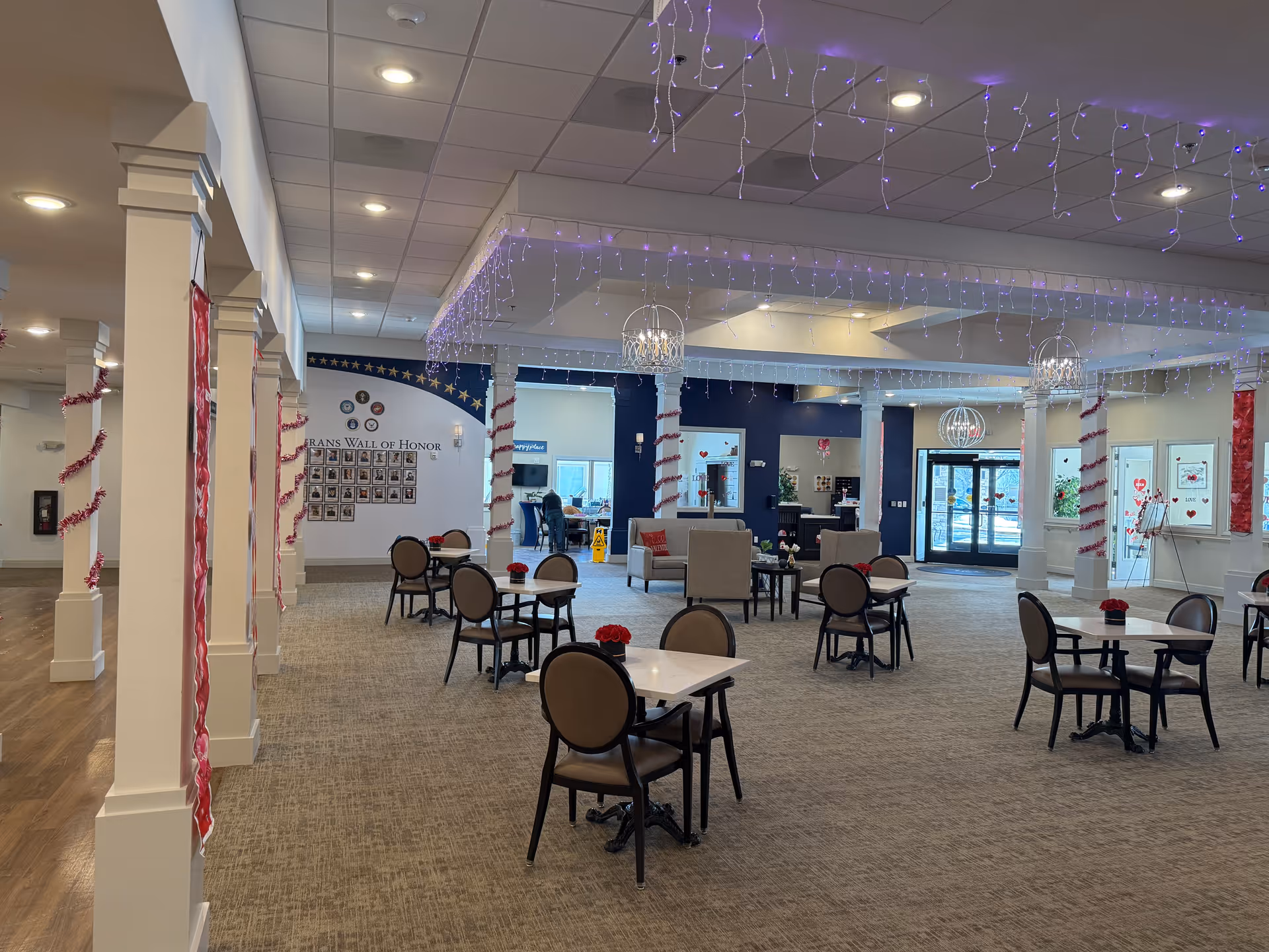 Spacious senior living facility common area with several small tables and chairs arranged on carpeted floor. The room features white columns decorated with red garlands, ceiling with recessed lighting and hanging string lights. Comfortable seating areas with armchairs and small tables are visible near the back. A wall labeled 'Veterans Wall of Honor' with photos is on the left side. Large windows and glass doors provide natural light.