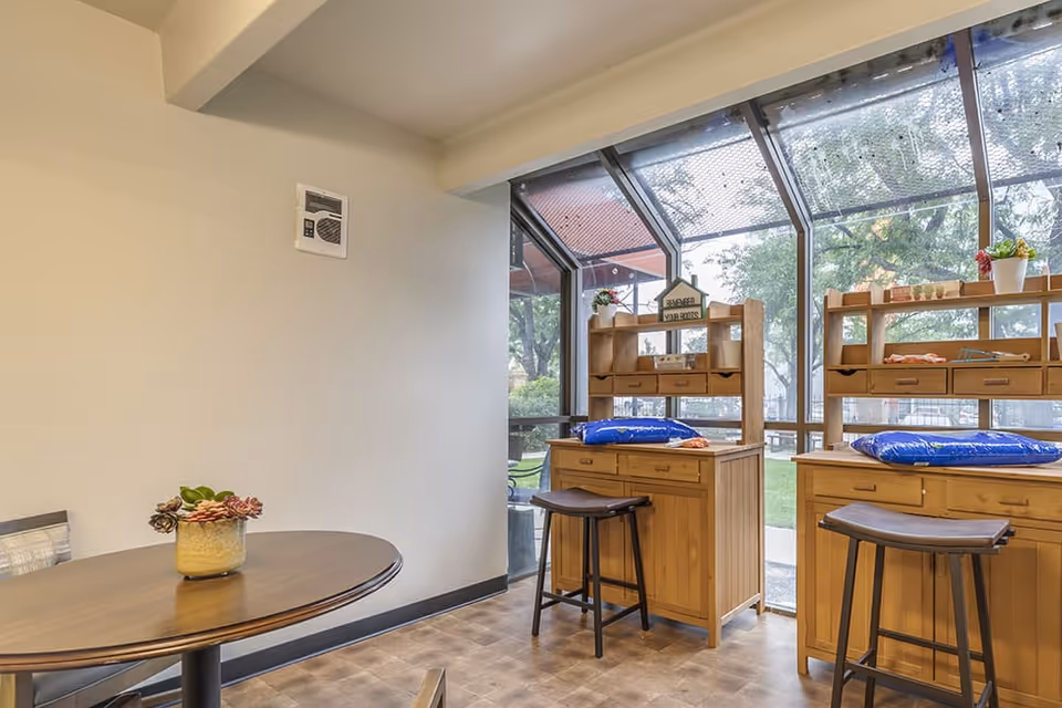 A bright room with large windows showing an outdoor view. Inside, there are two wooden desks with shelves and drawers, each accompanied by a tall stool. A small round table with a potted plant is also visible in the foreground. The floor has a patterned design and the walls are plain light-colored.
