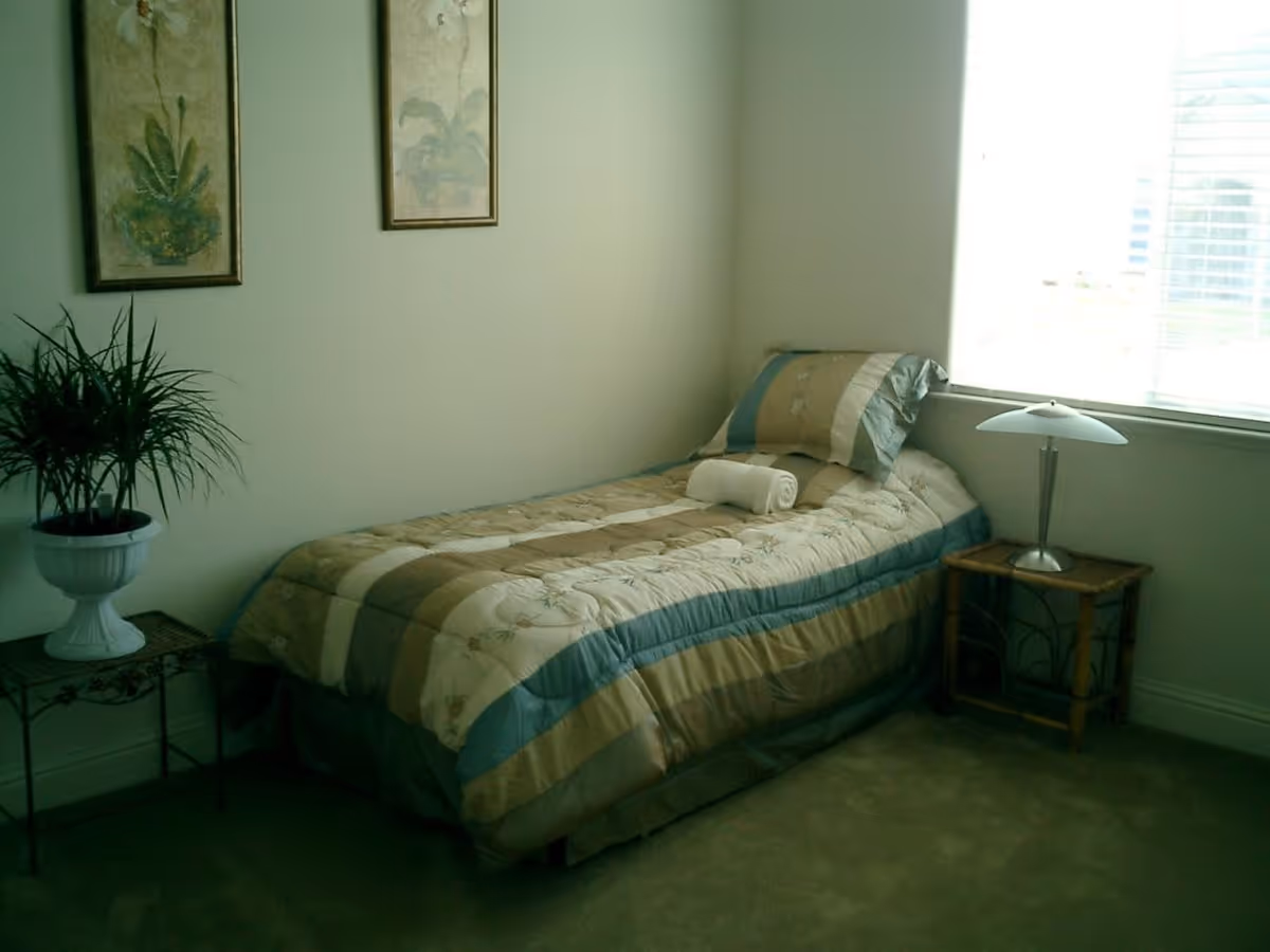 A small bedroom with a single bed covered in a beige, blue, and green striped comforter. A rolled white towel is placed on the bed. Next to the bed is a small wooden nightstand with a modern lamp. On the opposite side, there is a small table with a potted plant. Two framed botanical prints hang on the wall above the bed. A window with blinds allows natural light into the room.