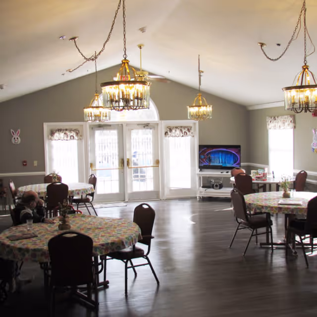 A spacious dining room with round tables covered in colorful tablecloths and surrounded by chairs. The room has large windows and glass doors letting in natural light, with chandeliers hanging from the ceiling. A television is placed on a stand against one wall, and a few decorative items are visible on the walls and tables.