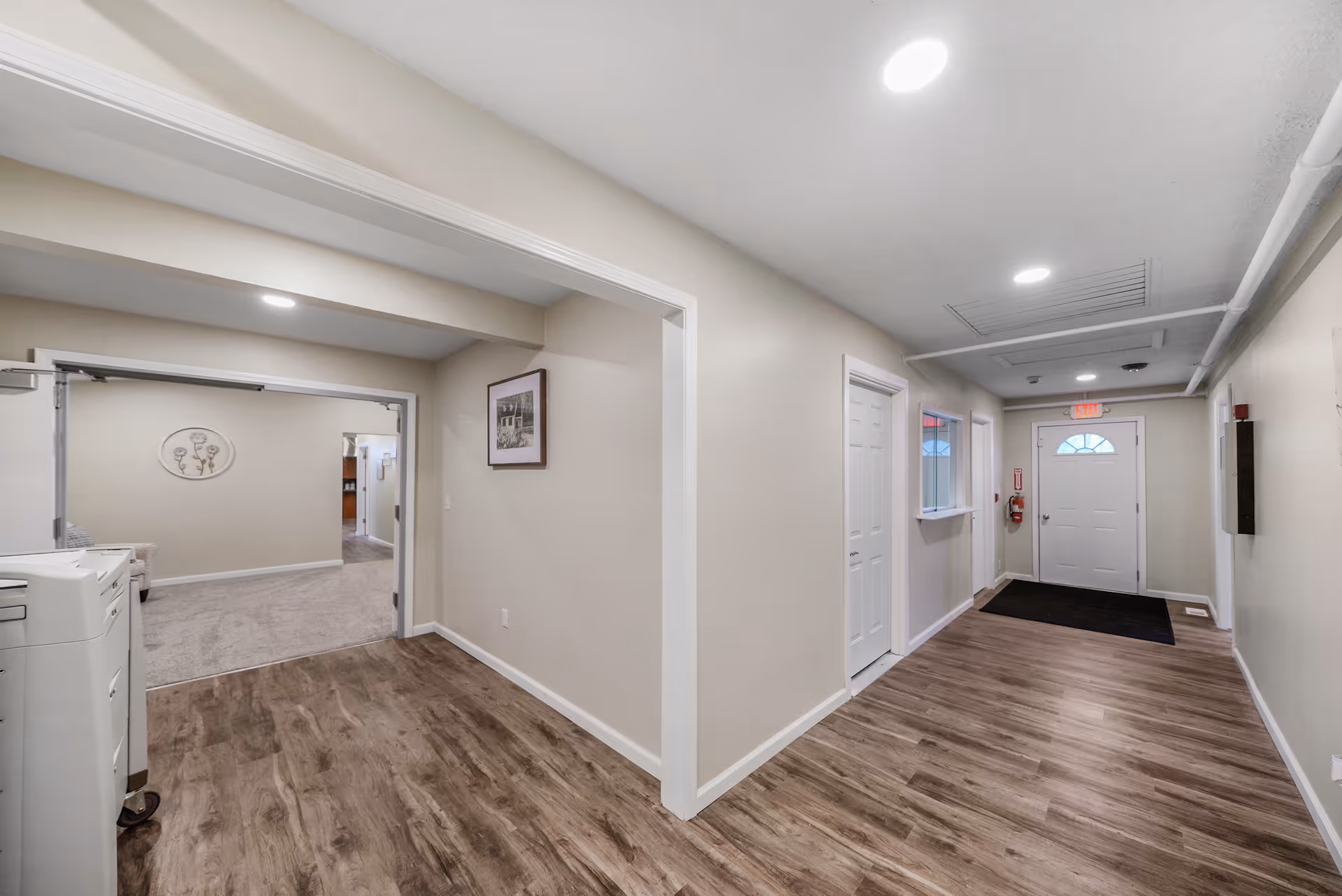Interior hallway of New Haven Assisted Living with wood flooring, beige walls, white doors, and ceiling lights. The hallway leads to a white door with a window and an exit sign above it. To the left, there is an open doorway leading to a carpeted room with wall decorations and furniture partially visible.