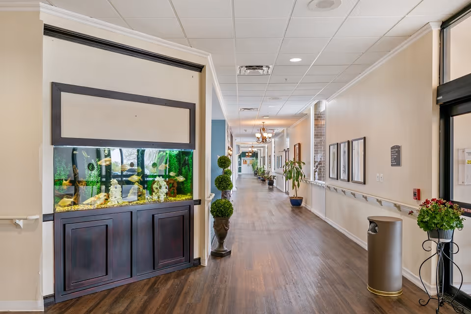 A long, well-lit hallway in a senior living facility with wooden flooring and beige walls. On the left side, there is a large built-in fish tank with various fish swimming inside. The hallway is decorated with potted plants and framed pictures on the walls. Handrails run along both sides of the hallway, and there is a trash can and a plant stand near the entrance on the right.