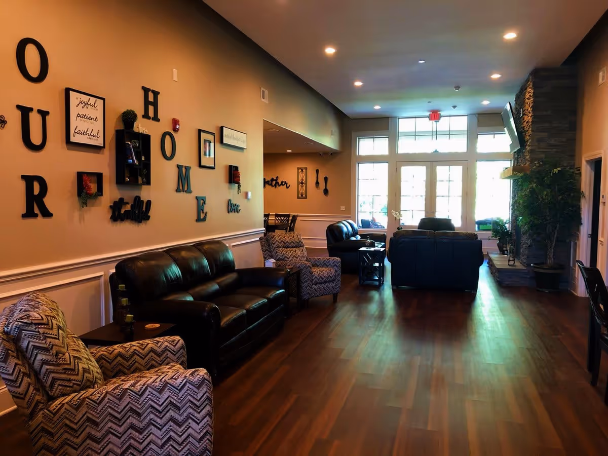 A cozy senior living facility common area with leather sofas, patterned armchairs, wooden flooring, and a stone fireplace. The wall is decorated with large letters spelling 'OUR HOME' and various framed inspirational quotes and small shelves with plants. Large windows and glass doors at the far end let in natural light.
