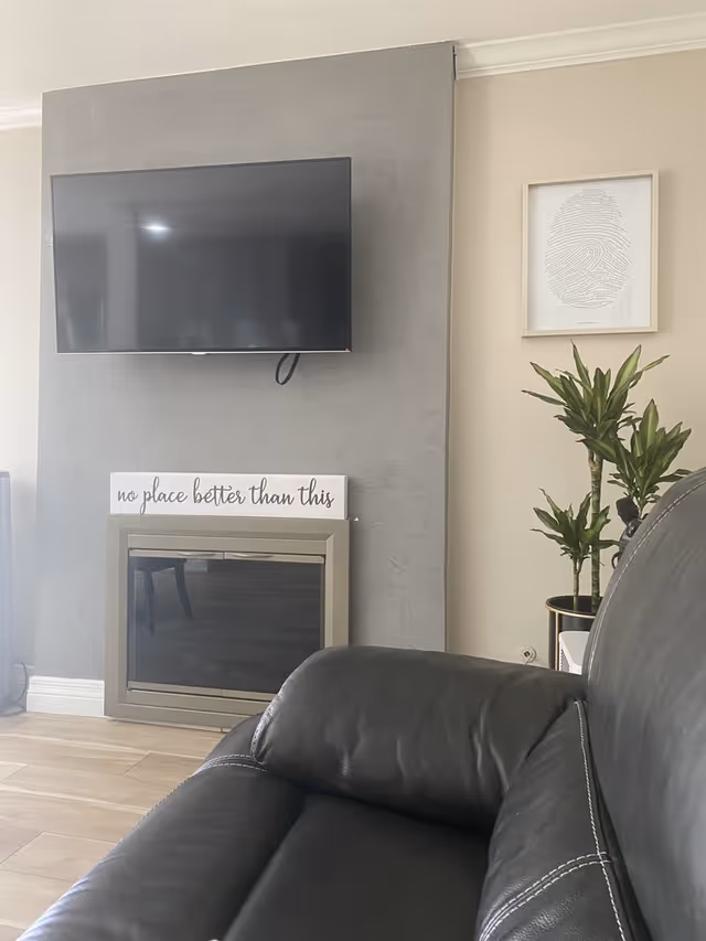 Wall-mounted TV above a decorative fireplace with a sign reading "no place better than this", a potted plant, and a black leather couch in the foreground of a living room.