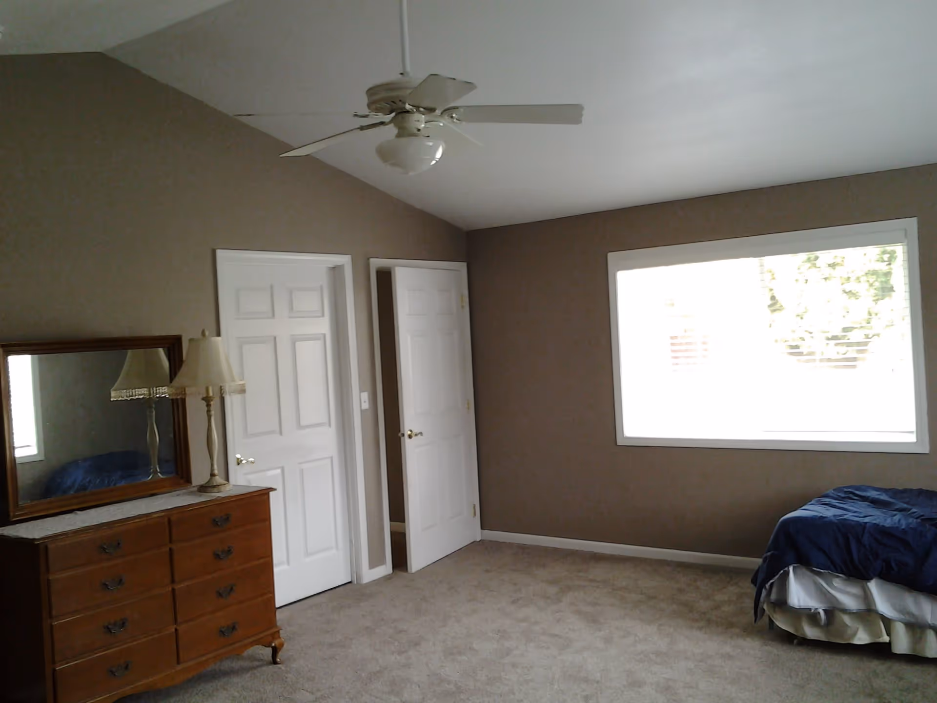 Spacious bedroom with a dresser and mirror on the left, a bed in the right corner, a large window, and a ceiling fan.