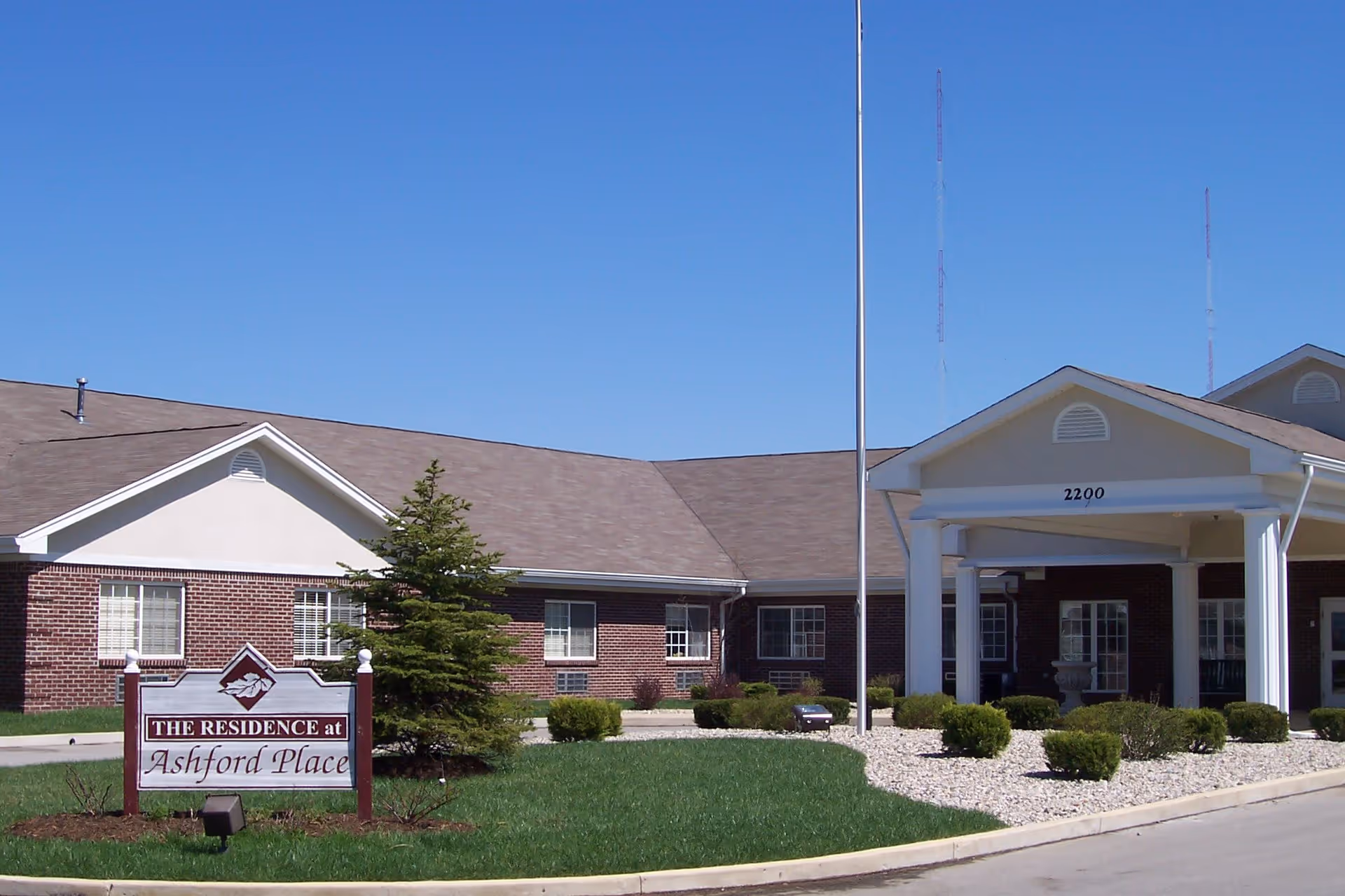 Exterior view of Ashford Place Health Campus showing a single-story brick building with a covered entrance supported by white columns. There is a sign in front that reads 'The Residence at Ashford Place' and a well-maintained lawn with bushes and a small tree under a clear blue sky.