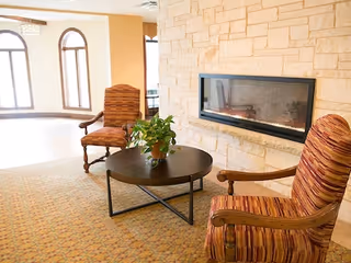 Two striped armchairs face a round coffee table with a potted plant in front of a built-in linear fireplace set in a stone wall, with arched windows in the background.