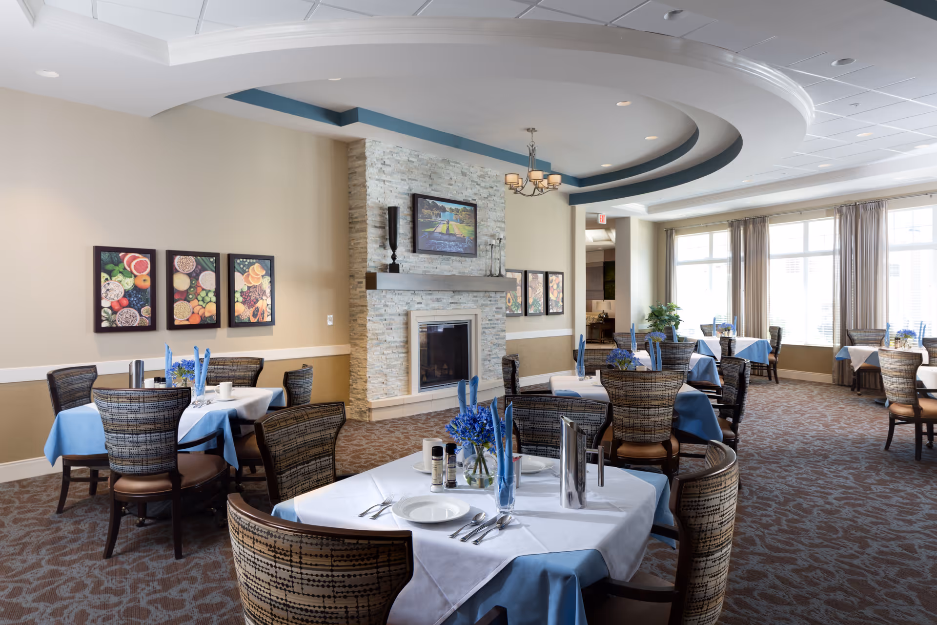 A bright dining room with several tables covered in white and blue tablecloths, each set with plates, silverware, and blue napkins. The room features a stone fireplace with a mantel and a framed picture above it. There are large windows with beige curtains allowing natural light to fill the space. The walls are decorated with colorful framed artwork of fruits and vegetables. The ceiling has a decorative circular design with a chandelier hanging in the center.