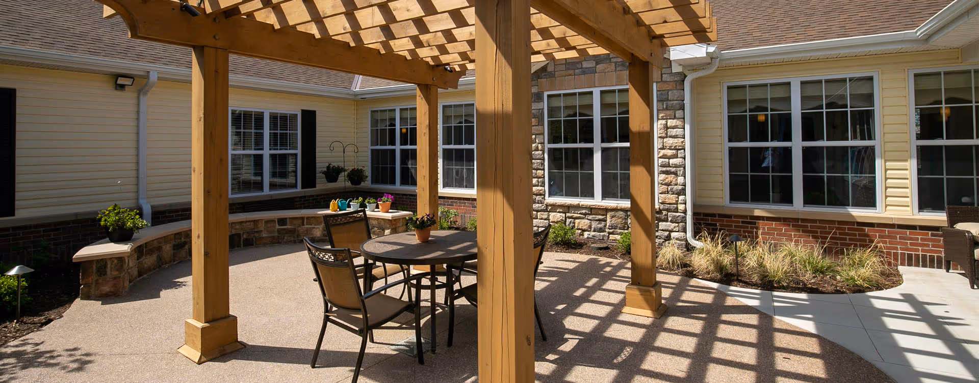 Outdoor patio area at Bickford of Saginaw Township featuring a wooden pergola casting shadows on a round table with four chairs. The patio is surrounded by a building with yellow siding, brick and stone accents, multiple windows, and some potted plants and landscaping.