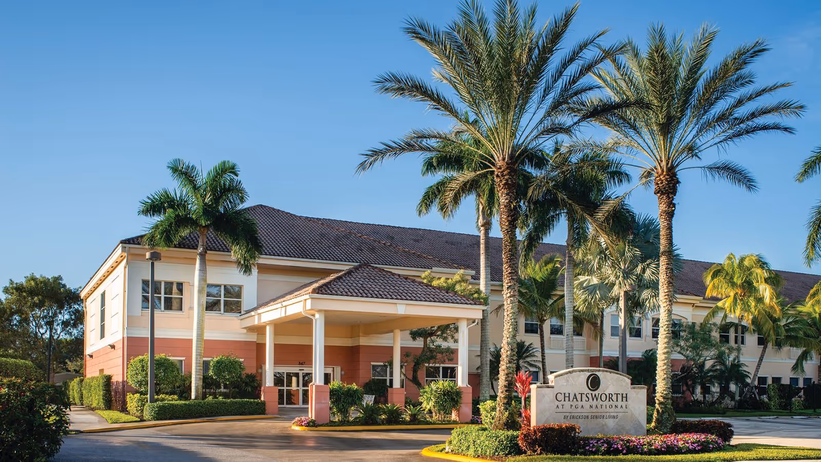 Exterior view of Chatsworth Assisted Living Community at PGA National, a two-story building with a covered entrance, surrounded by palm trees and well-maintained landscaping under a clear blue sky.