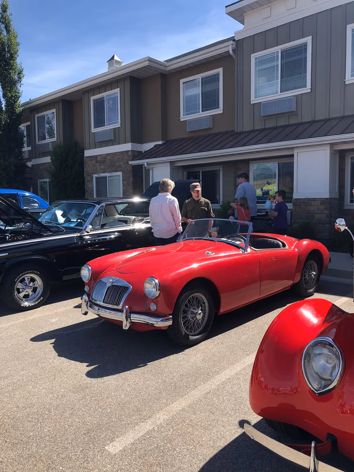 A red vintage convertible and other classic cars parked outside the front of a senior living building with people standing nearby.