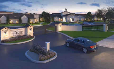 Evening view of the entrance to Wilshire Senior Living facility with a gated driveway, a parked black car, landscaped greenery, and the main building illuminated in the background under a partly cloudy sky.