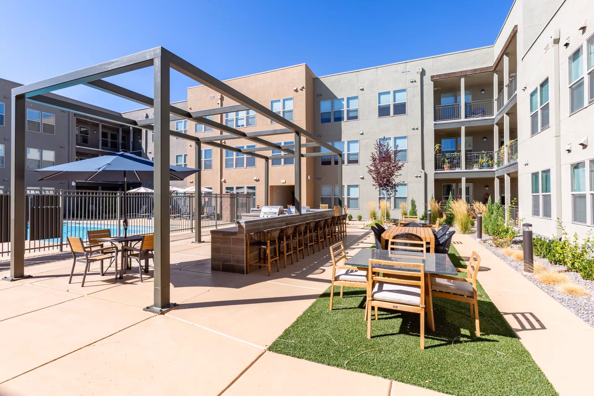 Outdoor courtyard with seating areas, umbrellas, a long bar counter and a fenced swimming pool next to a multi-story apartment building.