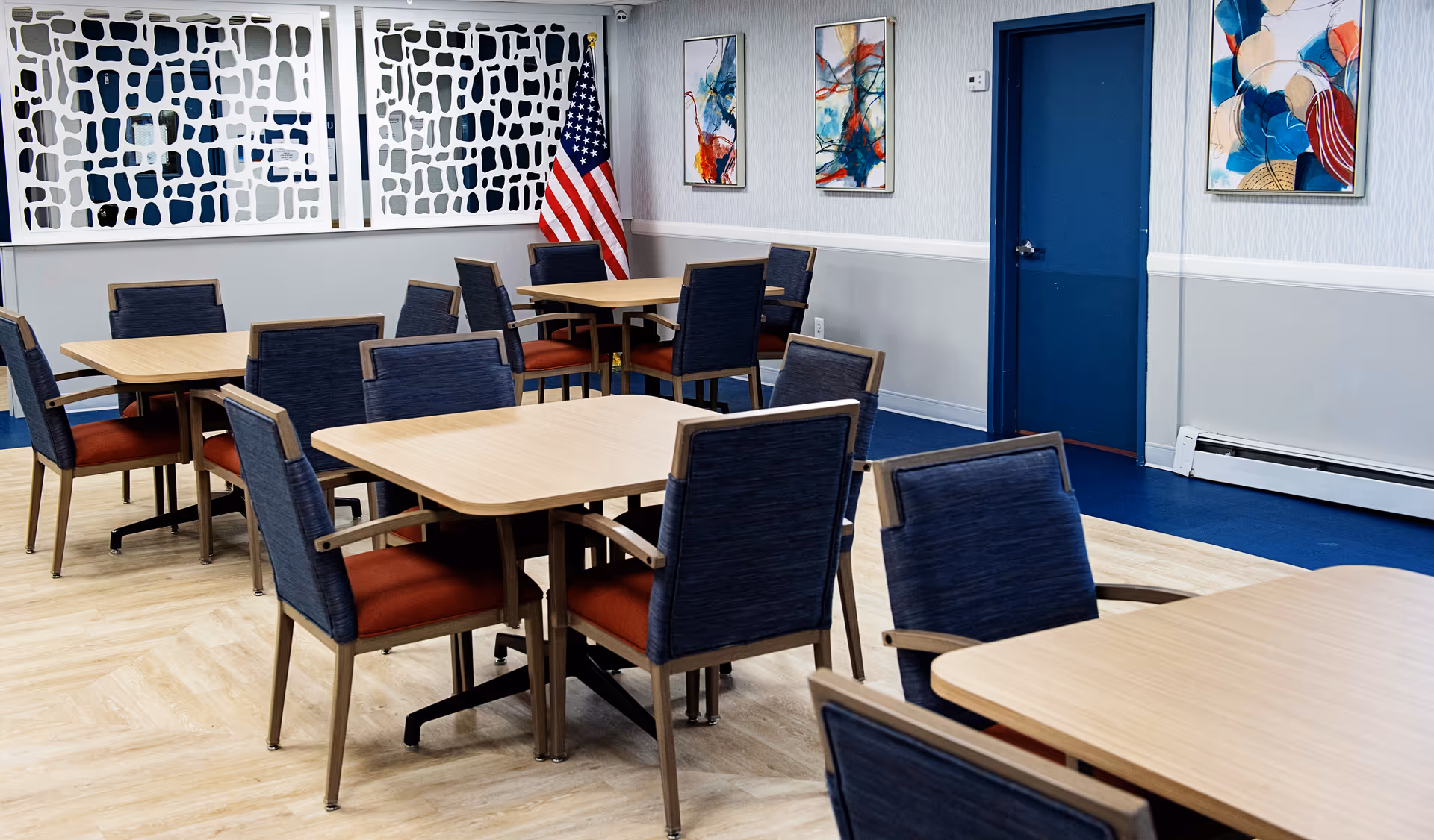 A dining room with several wooden tables and blue cushioned chairs with orange seats. The room has light wood flooring, a blue door, and abstract artwork on the walls. An American flag is positioned in the corner near decorative white panels.